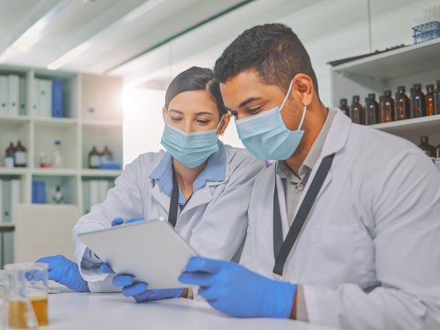 Two medical professionals look at a device in a laboratory. They are wearing white lab coats, blue gloves and blue face masks.