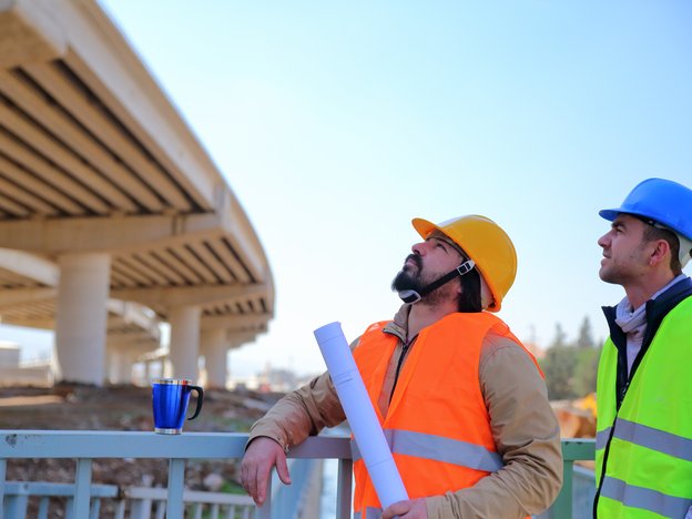 wo construction workers look up at a motorway bridge. They are wearing orange and green hi-vis vests.