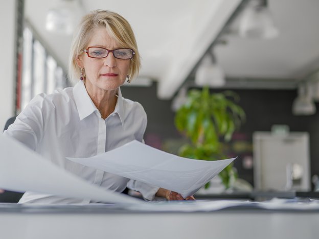 A woman organises pieces of paper at a desk. She sits in an open plan office.
