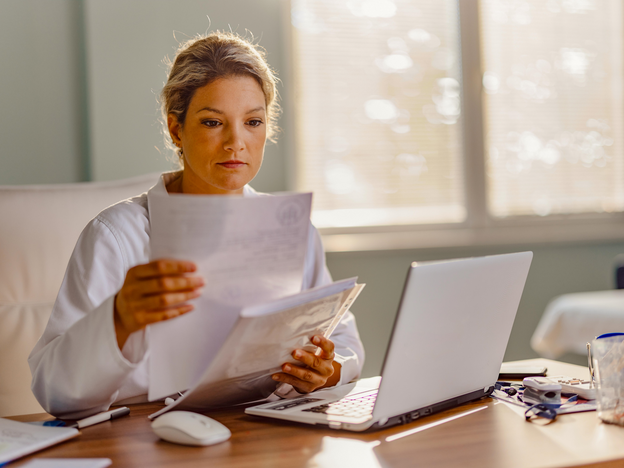 A woman reads documents at a desk. There is an open laptop and other office equipment on the desk.