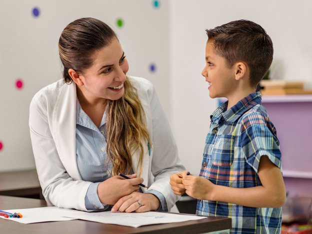 A female psychologist talks to a young boy in a clinic. They are leaning on a table and smiling. She is holding a pen and paper lies in front of them.