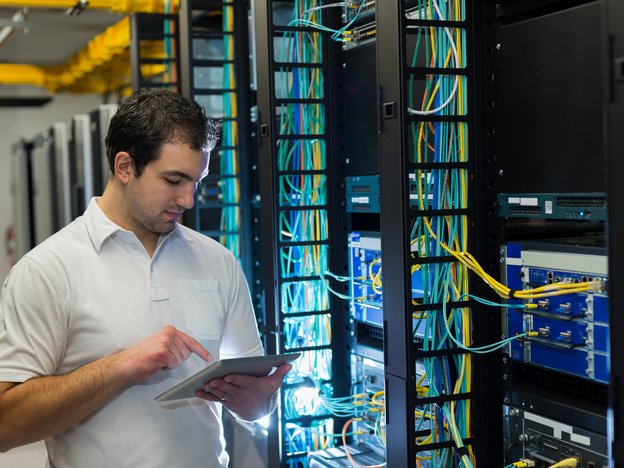 A man inspects a server bank and makes notes on a tablet.