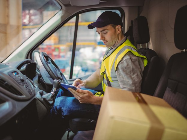 A male courier sits in the driver's seat of a delivery vehicle observing a device. A cardboard box is in the passenger seat.