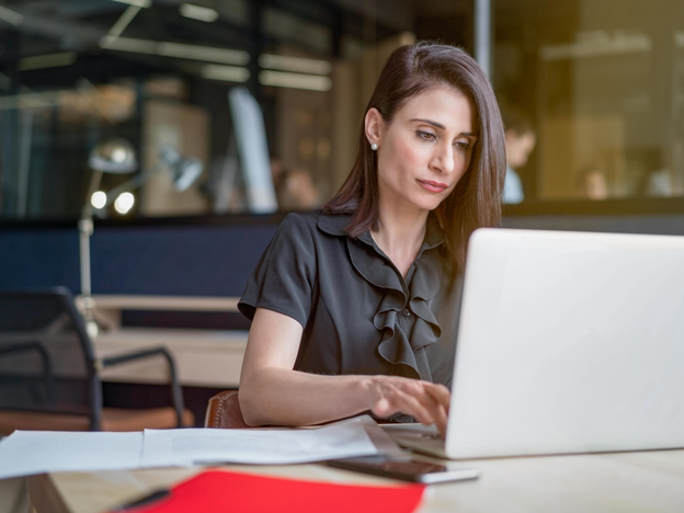 A woman types on an open laptop at a table. There are sheets of paper on the table.