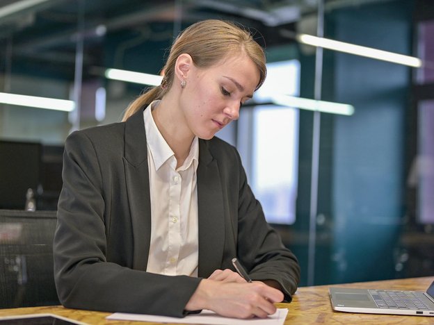 A young woman with a ponytail wears a black blazer and white shirt. She sits at a desk with a laptop and writes on a notepad with a pen.
