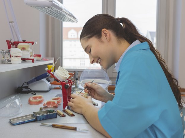 A dental technician smiles as she works on some teeth moulds sitting at a desk. She wears an aqua blue smock.