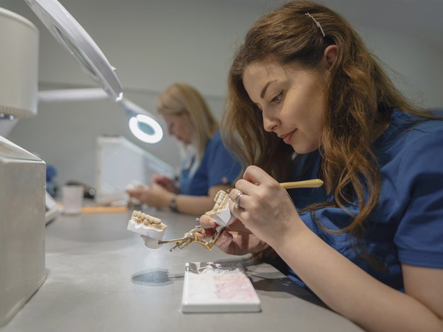 A female dental technician works on a set of teeth moulds under a microscopic light with another working woman next to her in the background. Both women wear a dark blue smock.