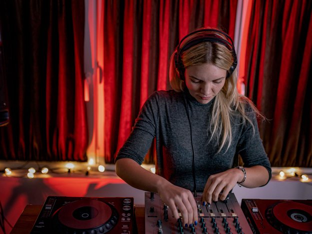 A female disc jockey wearing headphones mixes music using turntables. She is standing in a dark music venue.
