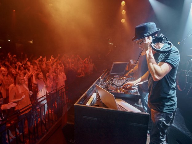 A male dj mixes music using turntables and stands in front of a crowd at a music venue. He is wearing a black hat and holds black headphones.