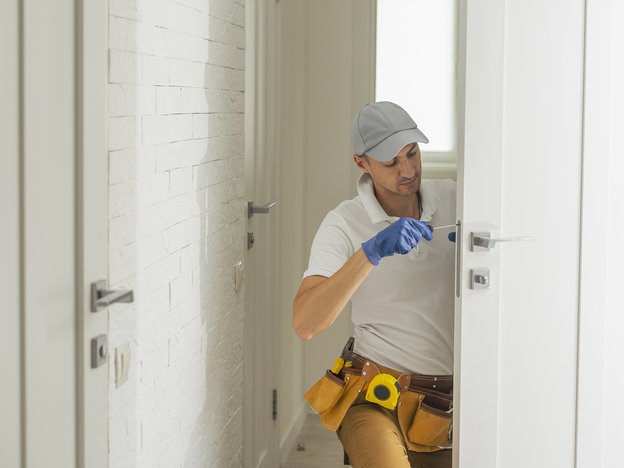 A man kneels on the ground and screws in a piece of metal on the side of a door. He wears a blue glove and a tool belt.