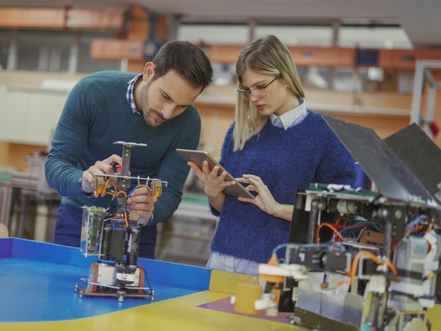A man and woman in a laboratory stand working on an electronics device. The woman wears a blue jersey while using a tablet and the man fixes the electronics model.