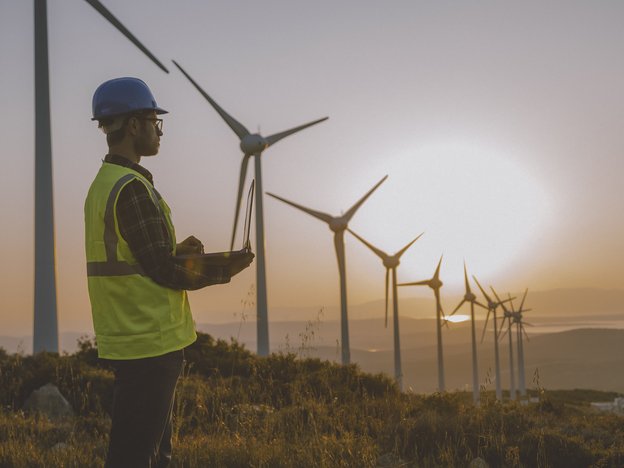 An man uses a laptop on a windfarm. He wears a hardhat, safety glasses and a hi-vis yellow vest.