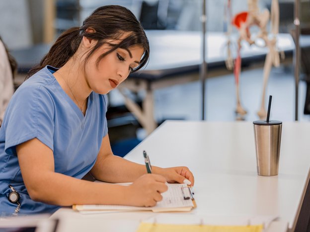 A woman writes on sheet of paper attached to a clipboard. She sits at a table and wears blue scrubs.