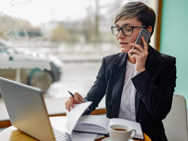 A woman looks at an open laptop and holds a mobile phone to her ear. She sits a table and there is a book, document and cup of coffee on the table.
