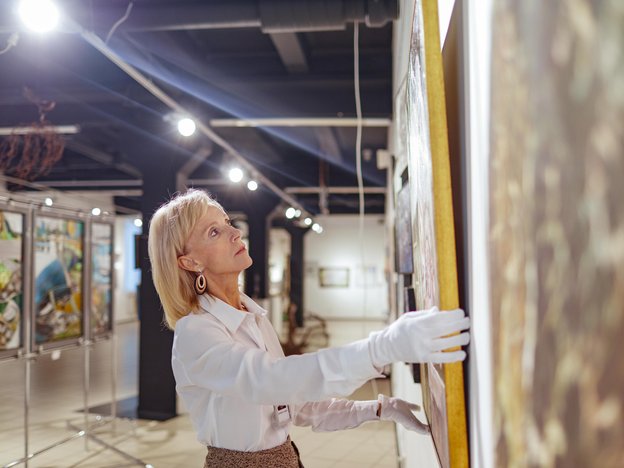 A womans places a painting on a wall. She wears white gloves and is inside an art gallery.