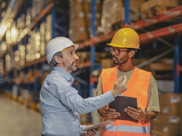 Two men interact with each other inside warehouse. One wears a high visibility orange vest and holds a clipboard. They both wear helmets.