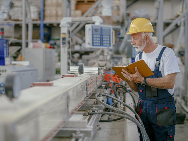 A man holds a brown notebook while he observes various machinery in a factory. He wears a yellow helmet and blue overalls.