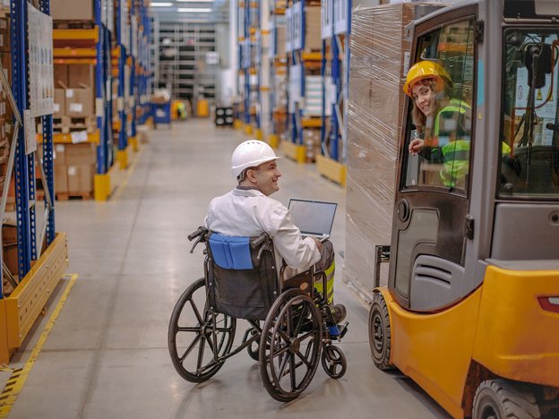 A man in a wheelchair smiles at a woman in an orange vehicle in a factory. He rests a laptop on his lap.