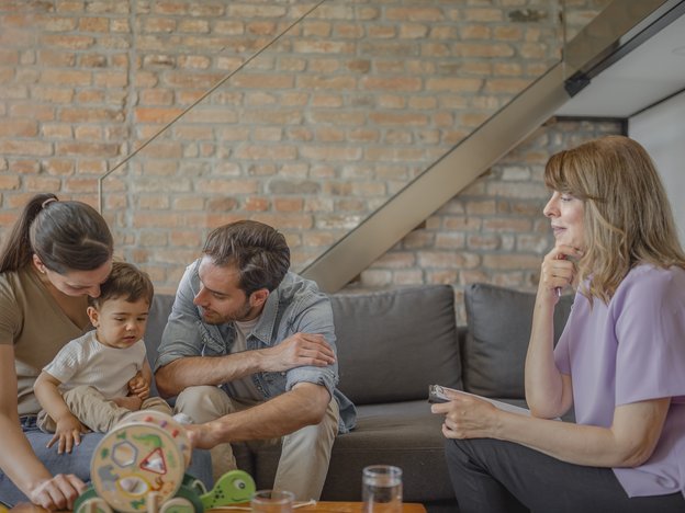 A man and woman sit on a couch with their young son sitting on the woman's lap. A woman counsellor sits opposite them reading from her notes during a counselling session.