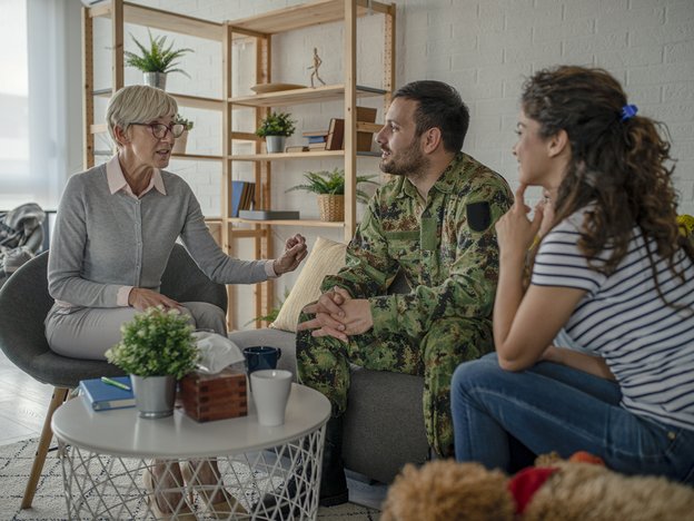 A couple sit on a couch interacting with a female counsellor during a counselling session. The counsellor wears a grey cardigan with a pink collared shirt. The man wears an army suit and the woman wears a blue and white striped t-shirt with blue jeans.