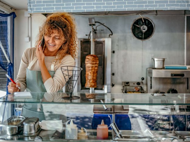 A woman takes a phone call behind a food service counter in a fast food restaurant. She is smiling as she notes down the order on a piece of paper with a pen.