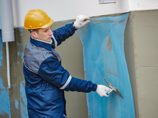 A man applies a blue sheet to a grey wall. he wears white gloves, blue overalls and a yellow helmet.