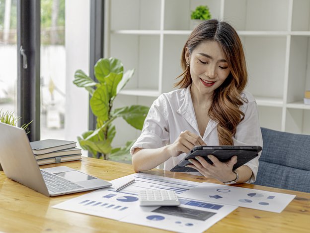 A woman holds and looks at her tablet. She sits at a desk in an office. There is an open laptop, calculator and documents on the desk.