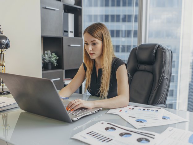 A woman types on an open laptop. She sits at a desk in an office. There are documents on the desk.