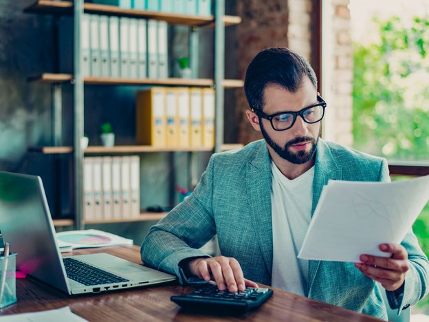 A man looks at a piece of paper at a work desk. An open laptop and calculator is in front of him.