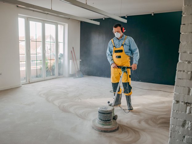 A man uses a machine to polish a concrete floor in a house. He is wearing a yellow apron and white facemask.