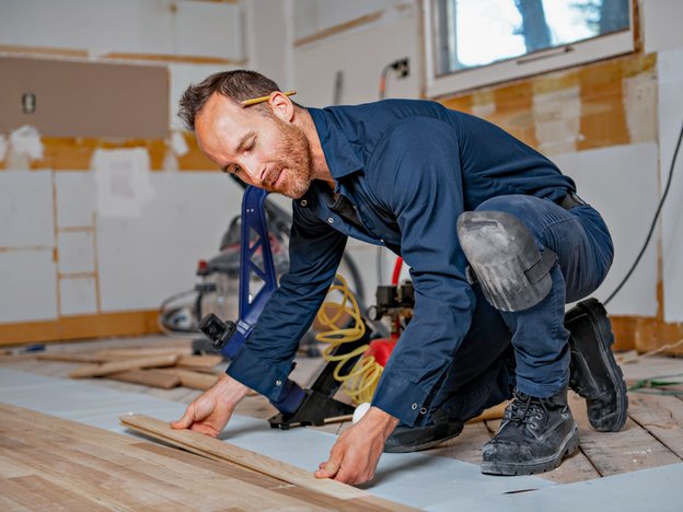 A man kneels down and places a long piece of wood onto a floor inside a home. He wears blue overalls.