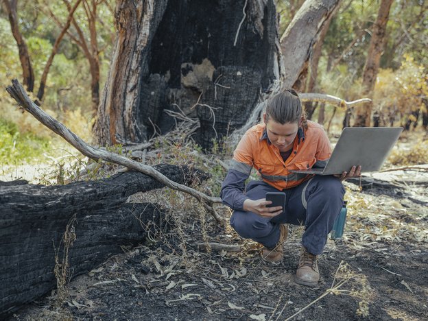A woman kneels beside a fallen tree. She holds a phone in hand and an open laptop in another. She wears high visibility orange and navy blue overalls