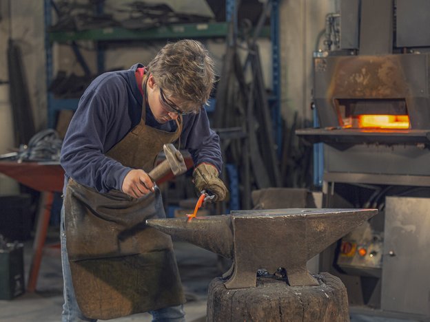 A man uses a chisel to forge some metal in a workshop. He wears a brown apron, brown glove and safety glasses. There is a lit fireplace behind him.
