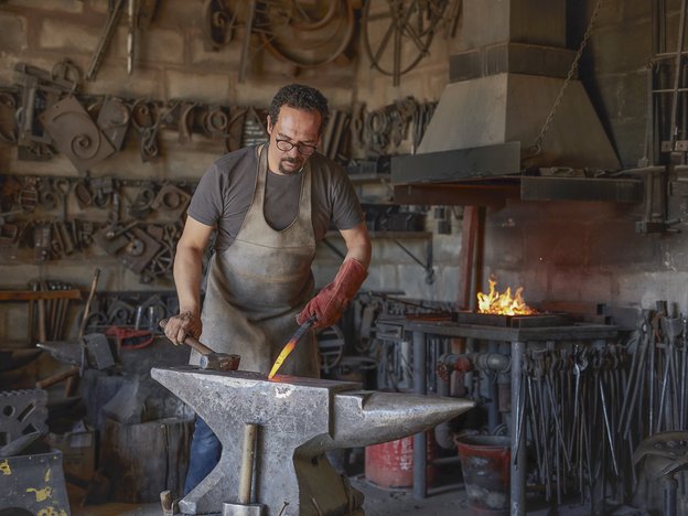 A man uses a chisel to forge some heated metal. He is wearing a brown apron and red glove. A lit fireplace is behind him.