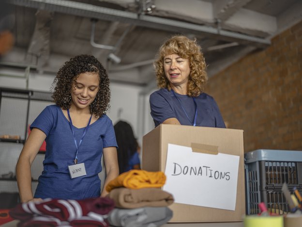 Two women fold clothes and place them into a cardboard box. The word 'Donations' on the box