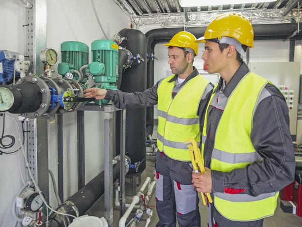 Two men look at a gas compressor. They both wear high visibility yellow vests, yellow helmets and grey overalls.