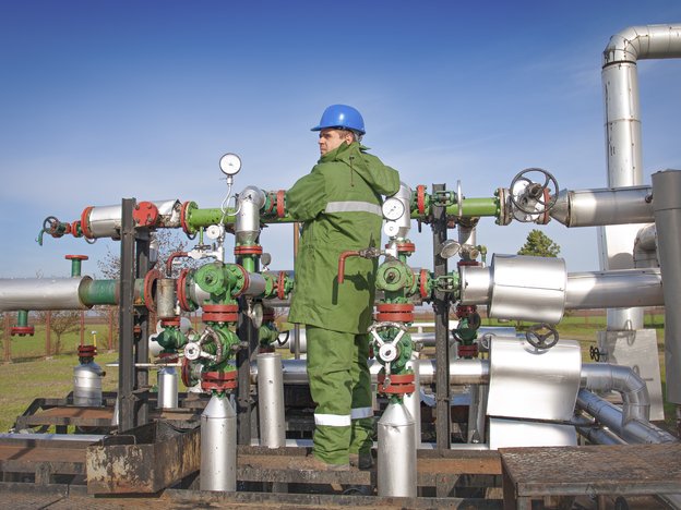 A man stands outside in front of gas pipes. He is wearing a green protective uniform and a blue helmet.