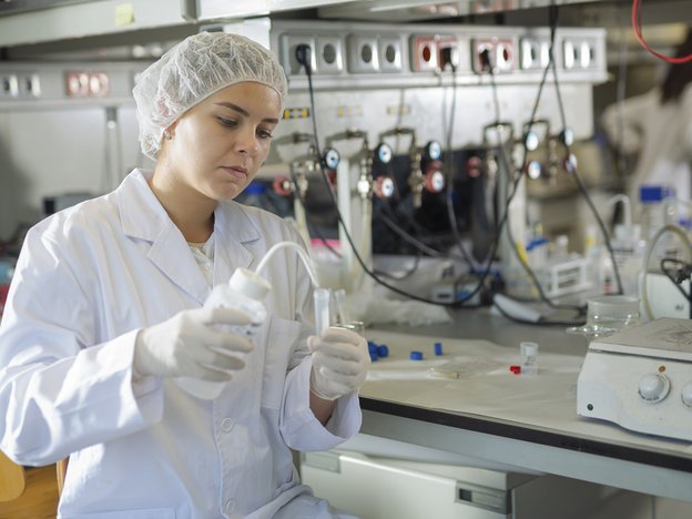 A woman transfers liquid from a bottle to a tube. She wears a hair net, lab coat and white plastic gloves. She is in a laboratory.