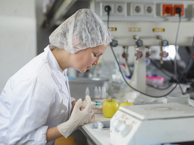 A woman injects a syringe into a pear. She wears a white lab coat, white gloves and a hair net.