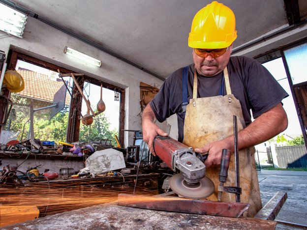 A man sharpens metal with a power tool. He wears a safety apron and a yellow helmet. He is inside a tool shed.