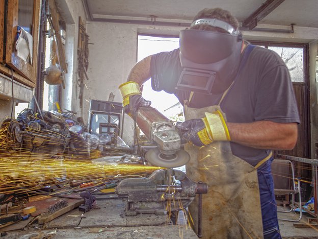 A man grinds metal that is sparking fire with a power tool. He wears yellow and blue gloves, blue overalls and a full facemask.
