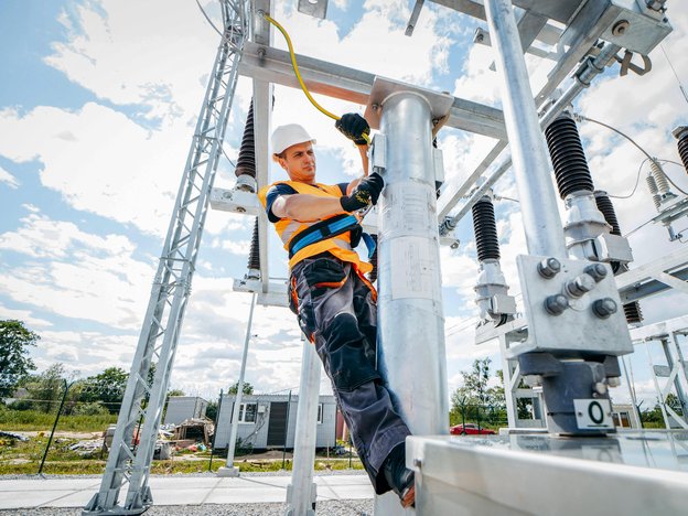 A man. attached to harness, applies a cord to a pole. He wears a white helmet, orange high visibility vest, black gloves and is at an electricity tower.