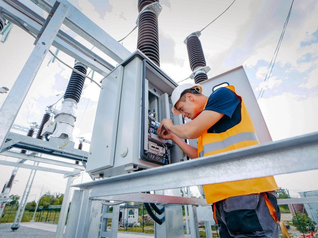A man inspects wires inside a tool box at a cell tower. He wears an orange high visibility house and protective white helmet.
