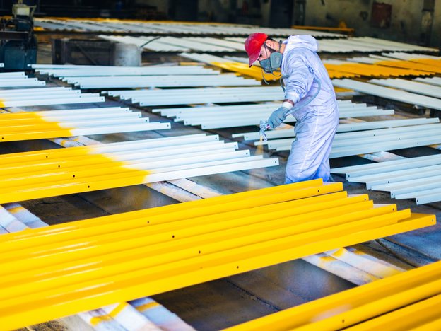 A man spray paints layers of steel yellow. He wears white overalls and a mask.