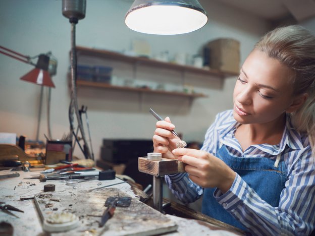A woman examines a ring with a pair of tweezers. She sits at a work desk.