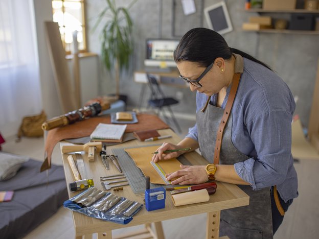 A woman measures leather on a desk using a ruler and various tools in a workshop. She wears a grey apron with brown straps.