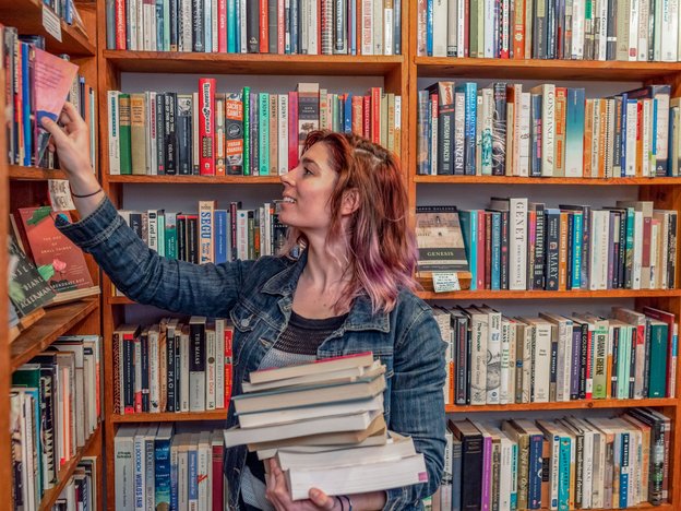 A female librarian takes or puts book back into a shelf in a library.  She is holding a stack of books.