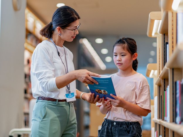 A female librarian shows a young girl a book. They are in a modern library.
