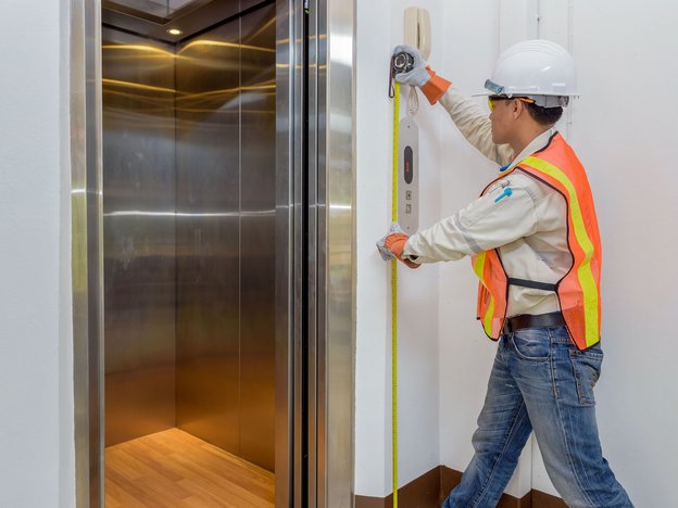 A man measures the length of a lift in a building with a tape measure. He wears a yellow and orange vest and a white helmet.