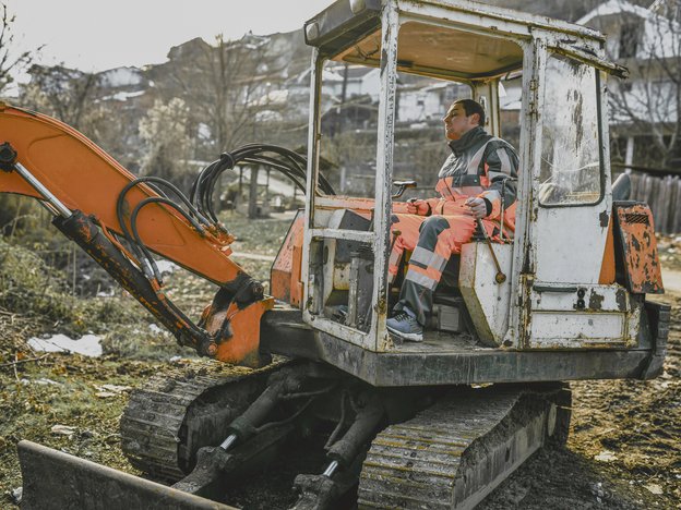 A man drives an orange and white excavator. He wears orange and black overalls.